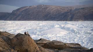 Two people sit on a rocky outcrop overlooking a vast glacier and ice-covered landscape, with rugged mountains rising beyond under a cloudy sky.