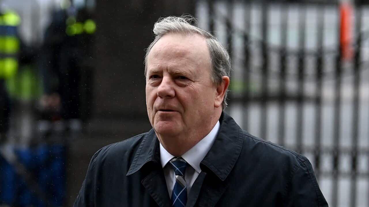 Peter Costello in a grey coat and suit walking. There are fences behind him.
