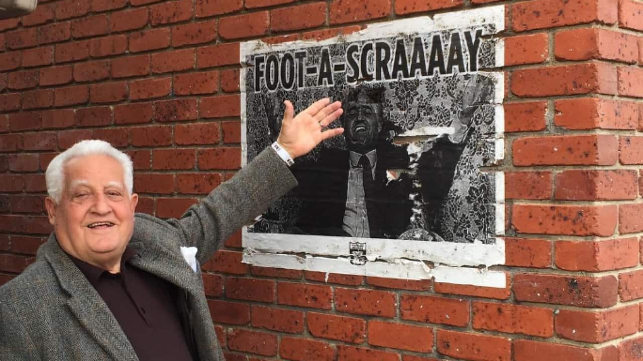 A man with white hair, wearing a grey jacket, is pointing at a black-and-white poster on a brick wall.