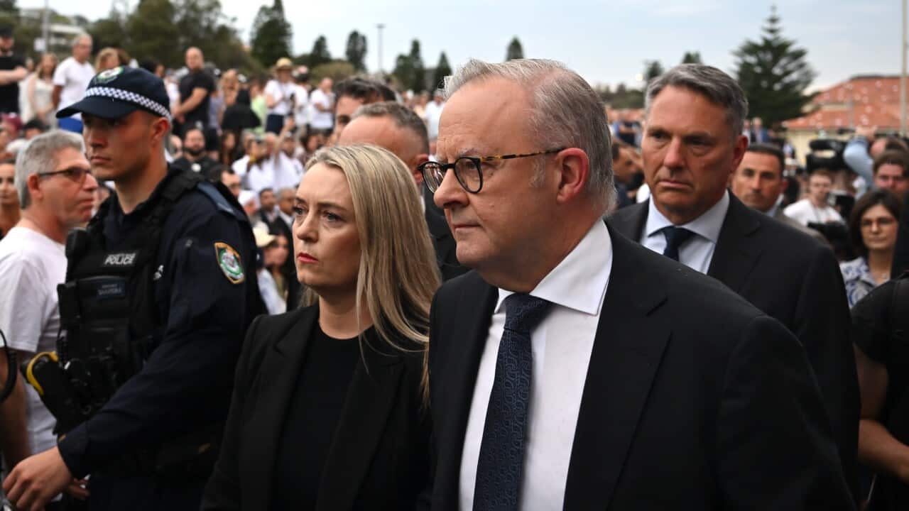 A man in a dark suit with glasses walks next to a woman with blonde hair in a crowd with a police officer in the background