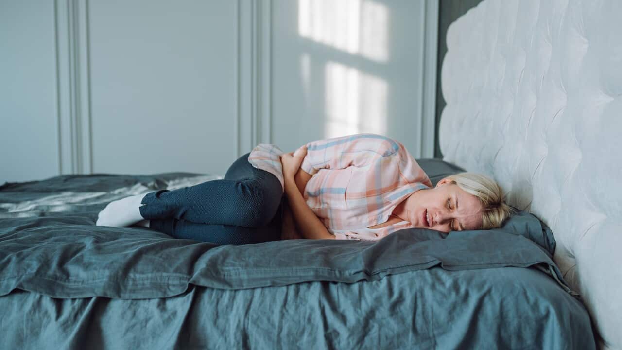 Menstruation period pain cramps. Blond woman grimacing, hugging herself on bed in light bedroom