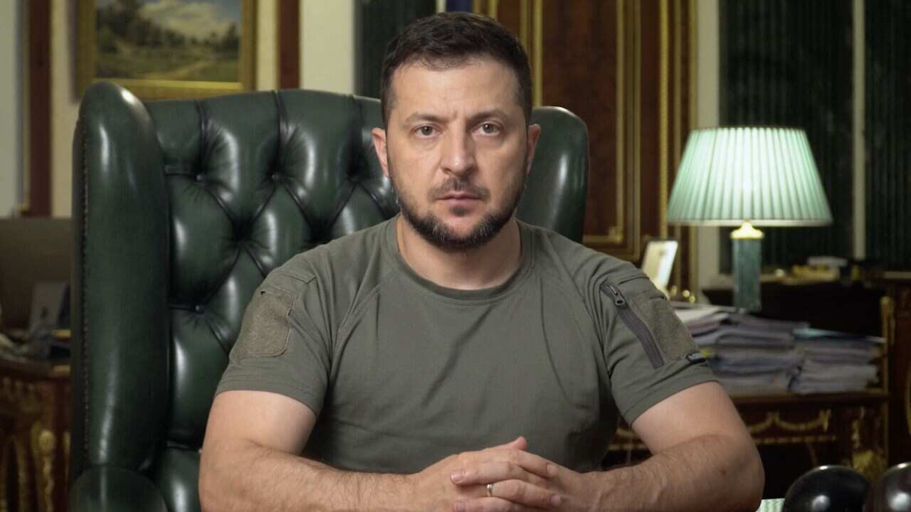 A man in a khaki t-shirt sits at a desk with his hands clasped in front of him