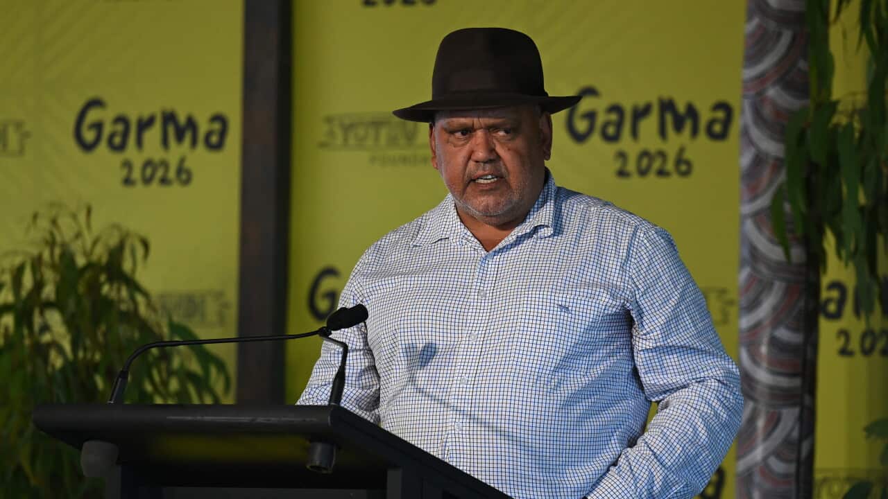A man wearing a black hat, checked long-sleeve shirt and jeans speaks at a podium during the Garma Festival 2026. Behind him are bright yellow backdrops displaying "Garma 2026" and the Yothu Yindi Foundation logo, with native plants framing the stage. He appears to be addressing an audience with a serious expression.