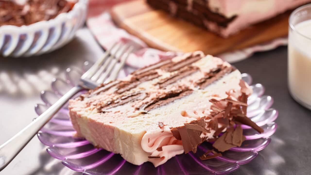 A retro scalloped purple glass plate holds a slice of ice-box xake, with chocolate biscuits sitting in cream and pink-coloured cream layers. A fork leans on the plate. Uncut cake sits on a serving board in the background.