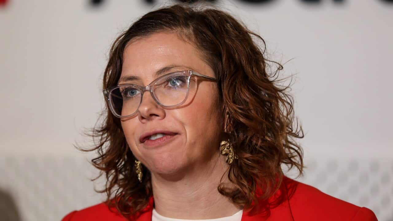 A woman with glasses wearing a black coat speaks at a press conference.