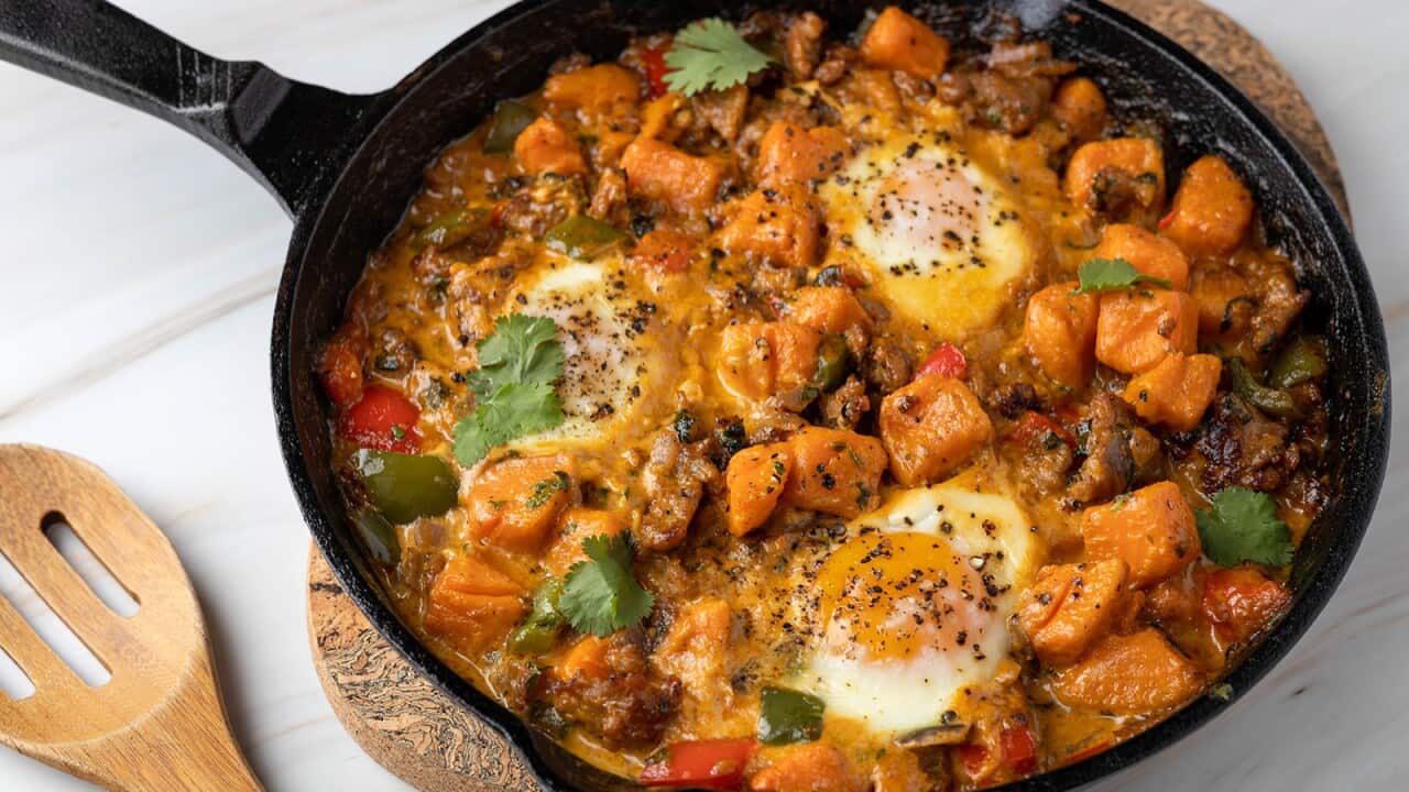 A cast iron skillet sits on a matt on a white surface with a slotted wooden spoon beside it. The pan has a stewish mixture of sweet potato, sausage, scattered herbs and two eggs that have been cooked in the mix.
