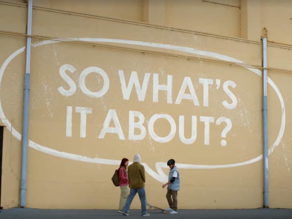 Three young people standing by a large wall which has a large speech bubble painted on it with the words 'so what's it about?' inside.