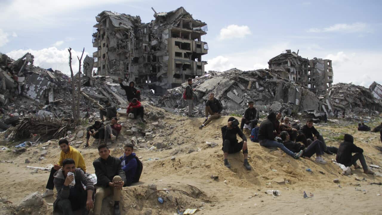 People sitting on the ground with rubble and damaged buildings in the background.