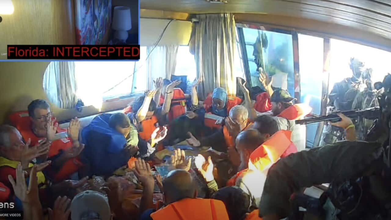 A group of people sitting inside a vessel, wearing lifejackets and raising their arms, as soldiers in uniforms point guns at them through a window.