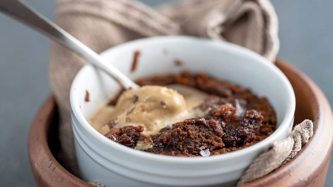 A white ramekin sits on a napkin in a larger brown ramekin. A delicious looking chocolate pudding, topped with melting ice-cream, is in the inner ramekin. A silver spoon sits in the pudding.