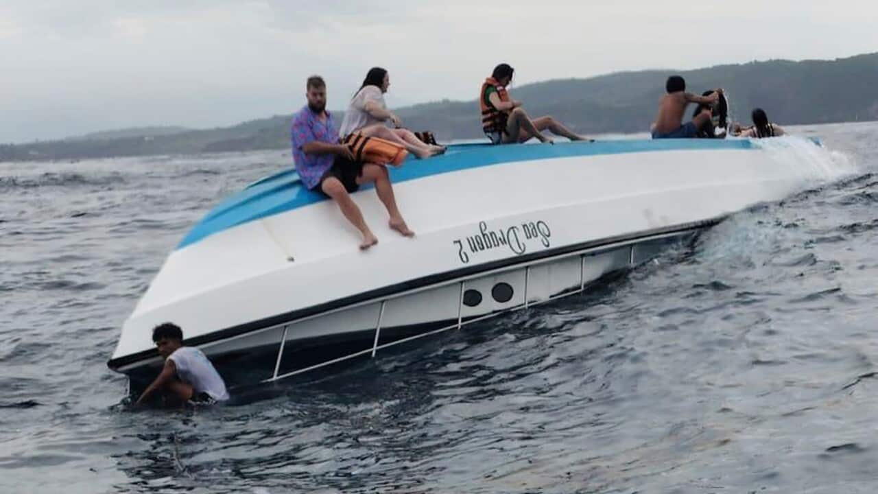 A partially capsized speedboat with people on top of it and in the water. Some individuals are holding onto the boat or swimming.