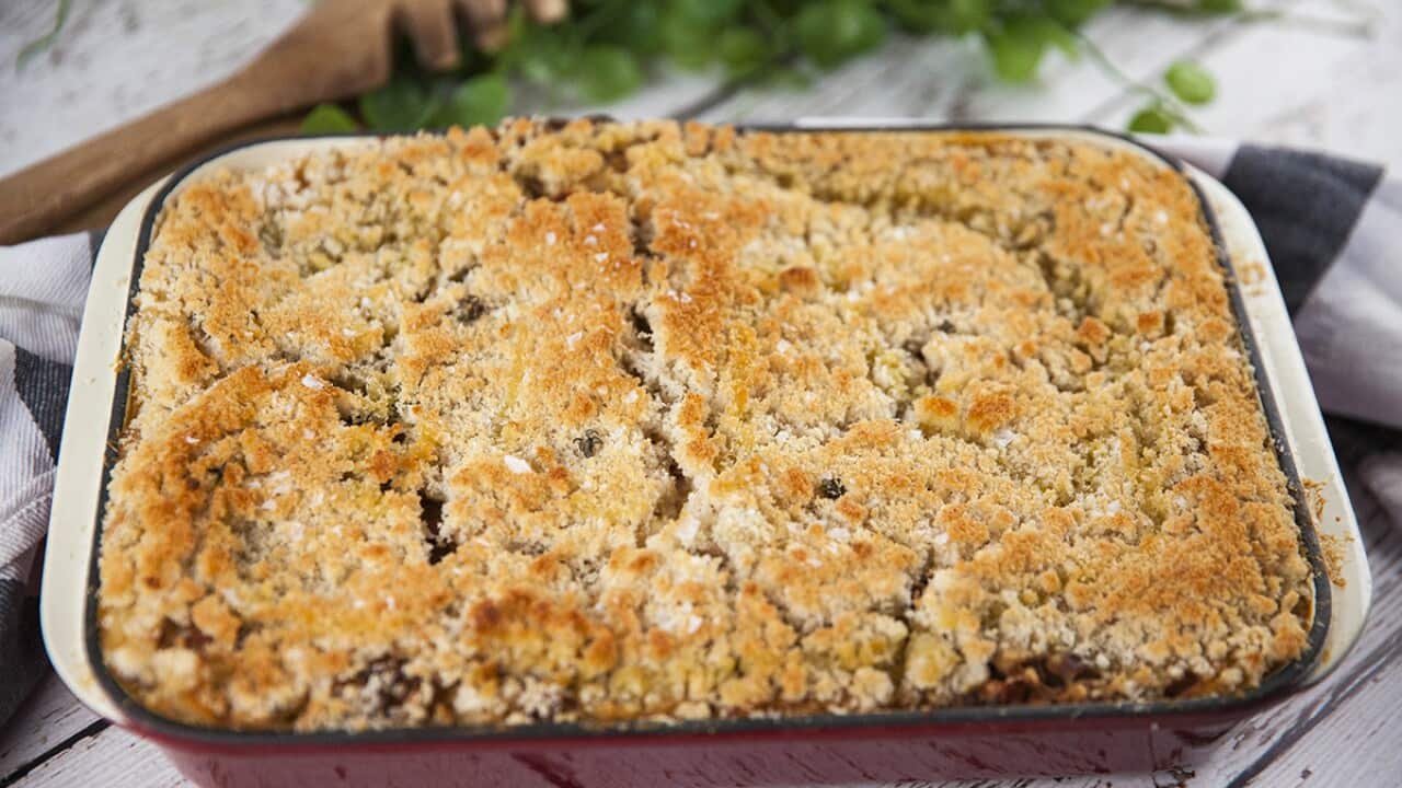 A deep red cast iron baking dish sitting on a white wooden surface is seen from overhead. It has a baked filling, topped with golden breadcrumbs.