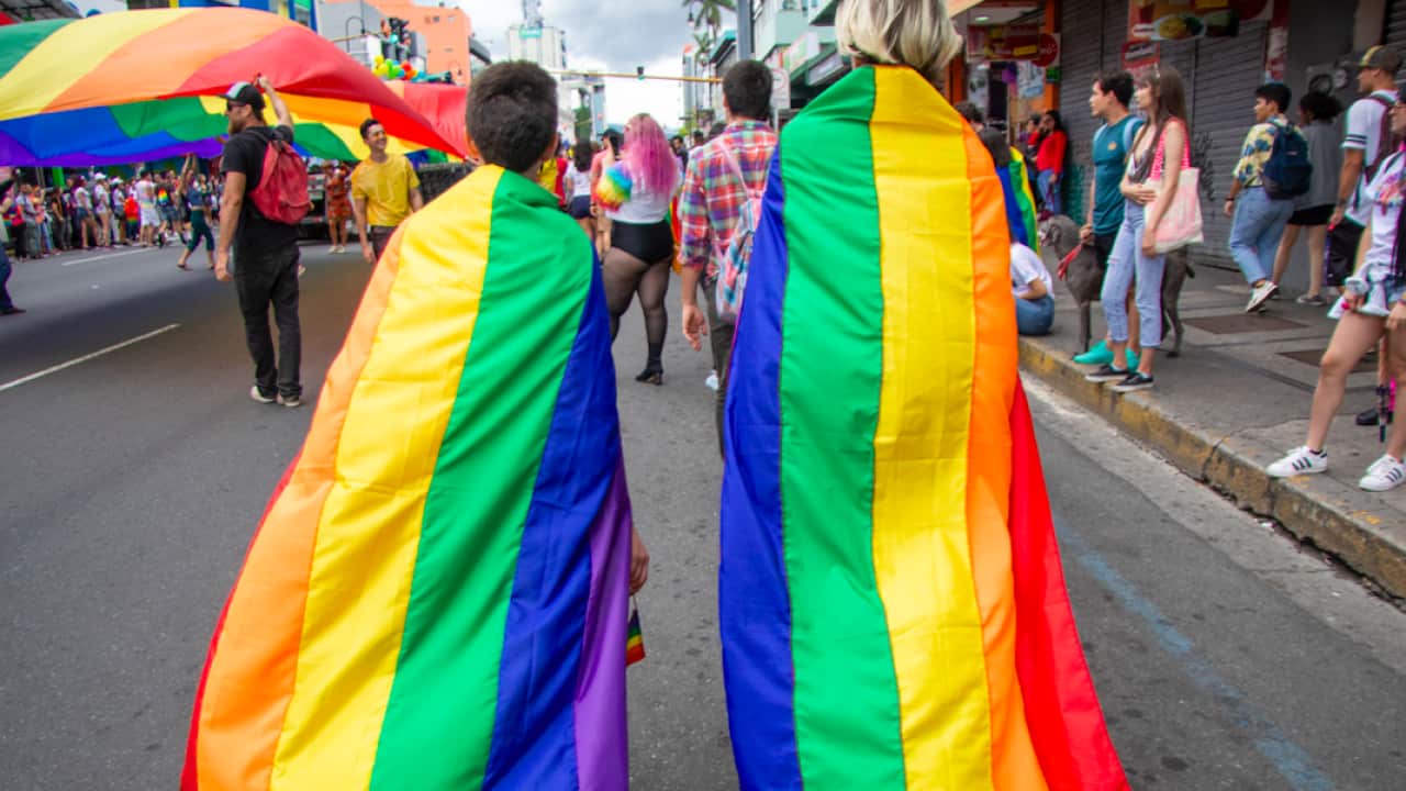 Two persons walk down a road wearing rainbow flags as capes.