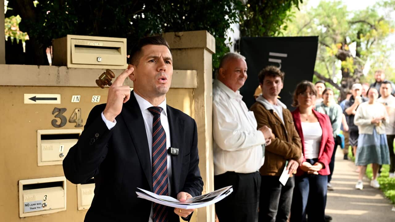 A man in a suit and tie in front of a residential property raises his finger while holding a small wooden hammer in his right hand and documents in his left hand, with people standing in the background