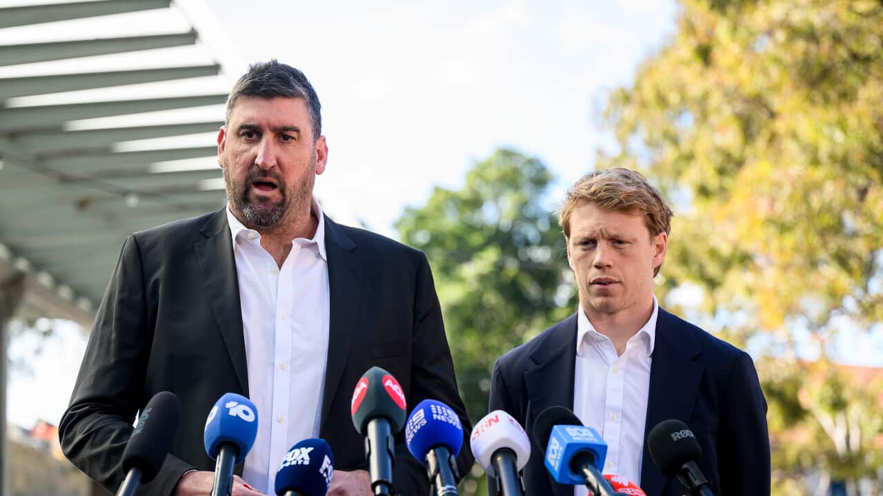 A man in a black suit and another in a navy blue suit stand before microphones at a press conference.