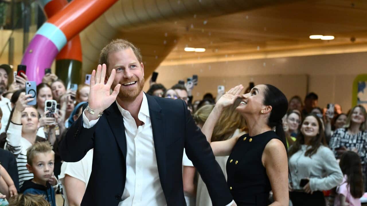 Prince Harry and Meghan, the Duke and Duchess of Sussex greet children during their visit to the Royal Children’s Hospital in Melbourne. Harry wears a black suit with white shirt. Meghan wears a dark sleeveless dress with gold buttons.