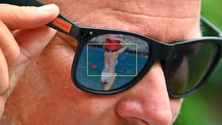Close-up of smart glasses with a built-in camera and recording indicator, showing a framed view in the lens reflection of a woman standing by the water.