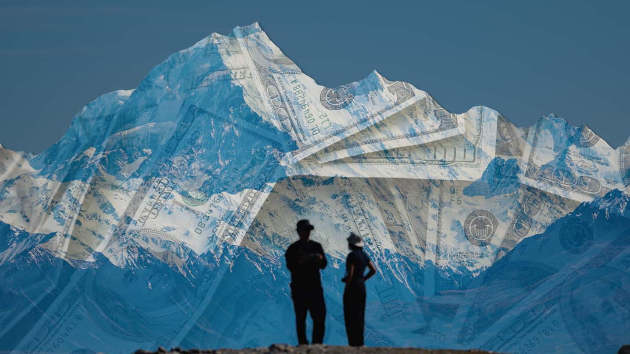 Two shadowed figures stand in front of a large mountain. The mountain is covered in translucent US bills.