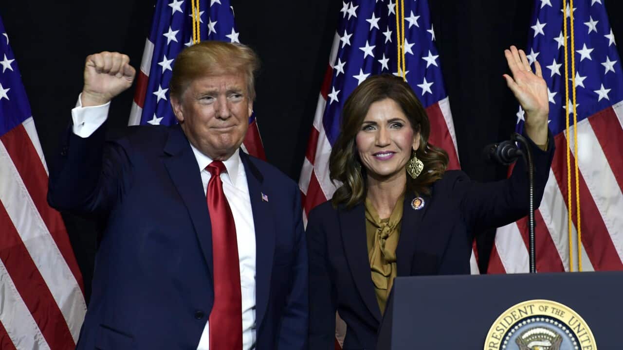 Donald Trump and Kristi Noems wave at a podium with American flags in the background