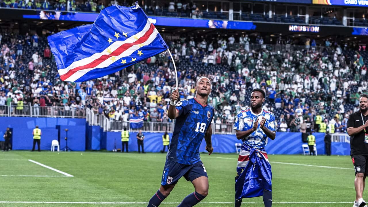 A Cape Verde player waves the national flag while walking across the pitch after the match, as a teammate applauds nearby. Fans in the stands celebrate in the background.