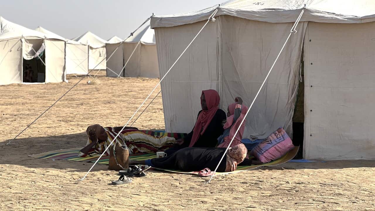 Three people rest outside a large white tent, sitting and lying on a mat on the sandy ground, with more tents visible in the background under a bright, sunny sky.