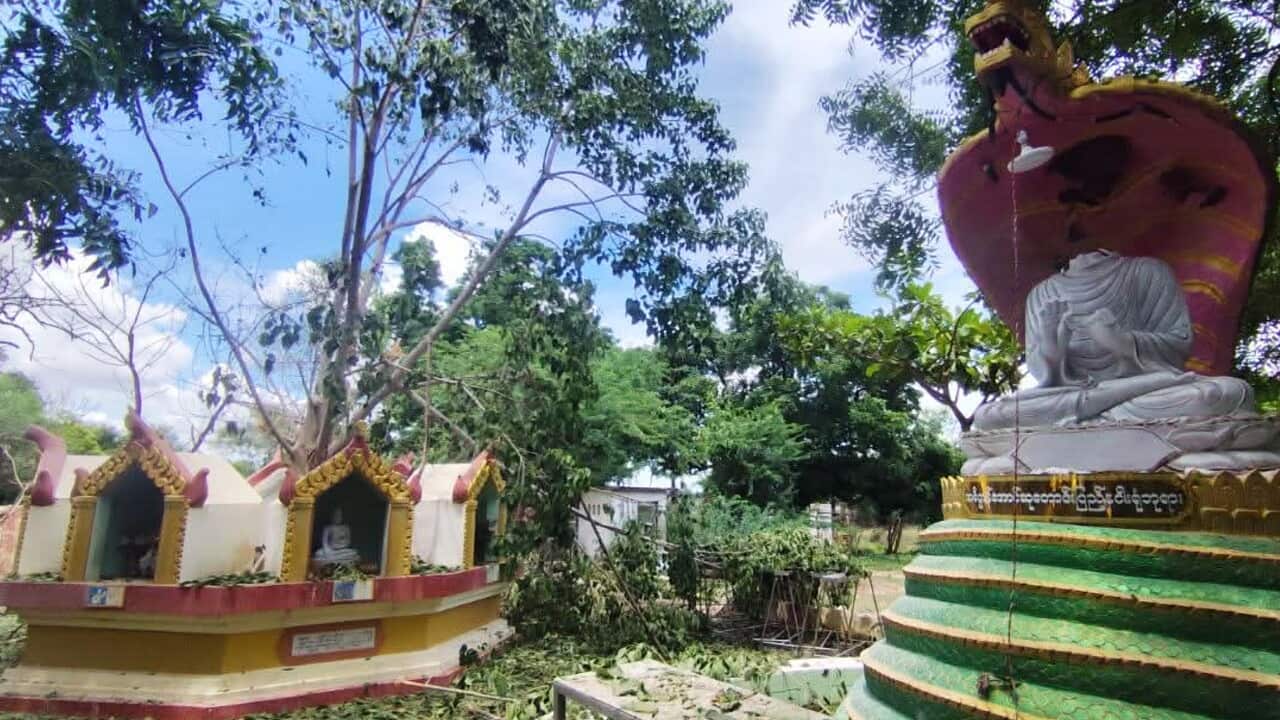 A Buddhist monastery complex in Myanmar, with a large seated Buddha statue on the right and smaller shrine structures on the left, surrounded by trees and scattered debris.