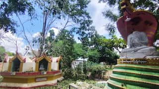 A Buddhist monastery complex in Myanmar, with a large seated Buddha statue on the right and smaller shrine structures on the left, surrounded by trees and scattered debris.