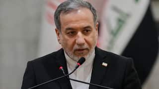 A middle-aged man with short grey hair and a beard, wearing a black suit and white shirt with a small lapel pin, is captured in a medium close-up as he speaks into two microphones against a blurred flag background.