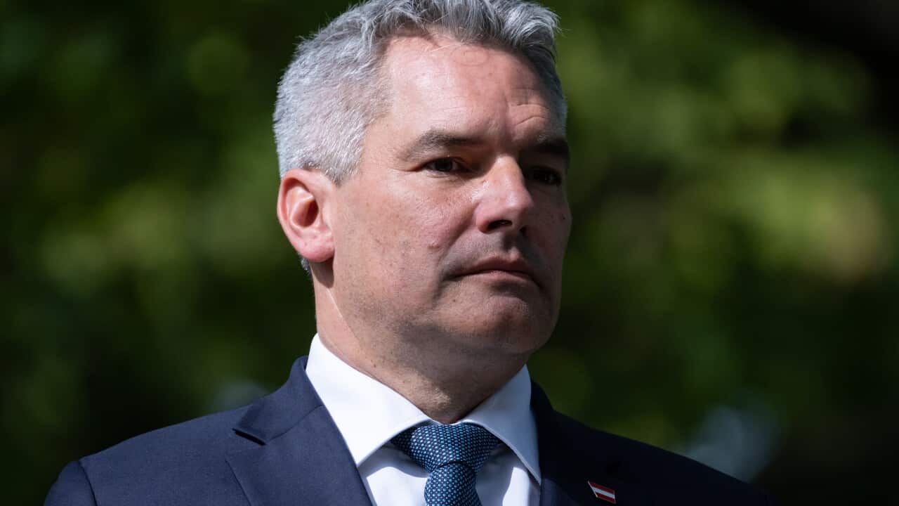 A man in a blue suit, white shirt and blue tie standing in front of trees.
