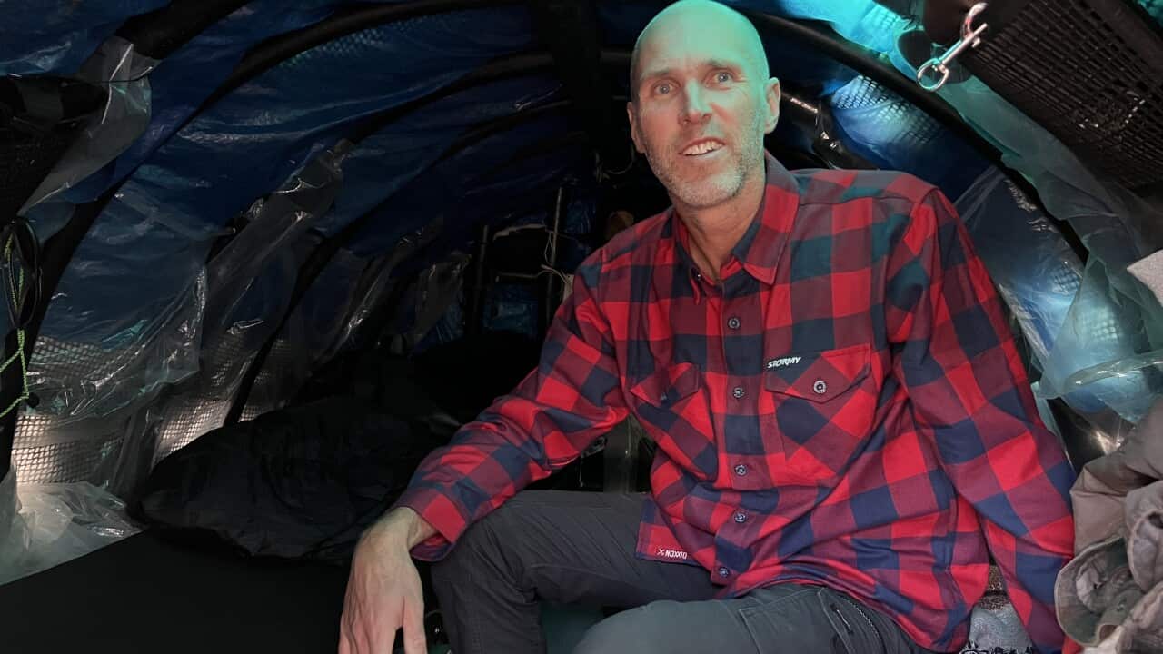 A man wearing a red and black check flannelette shirt and dark trousers sits in a boat cabin