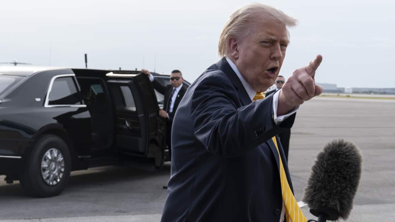 Donald Trump stands on an airport tarmac beside a black SUV, pointing and speaking toward reporters. A large microphone is visible in the foreground, while security personnel stand near the open car door in the background.