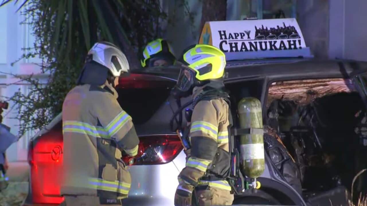 Firefighters inspect a burnt-out car outside a building, with a “Happy Chanukah” sign visible on the roof.