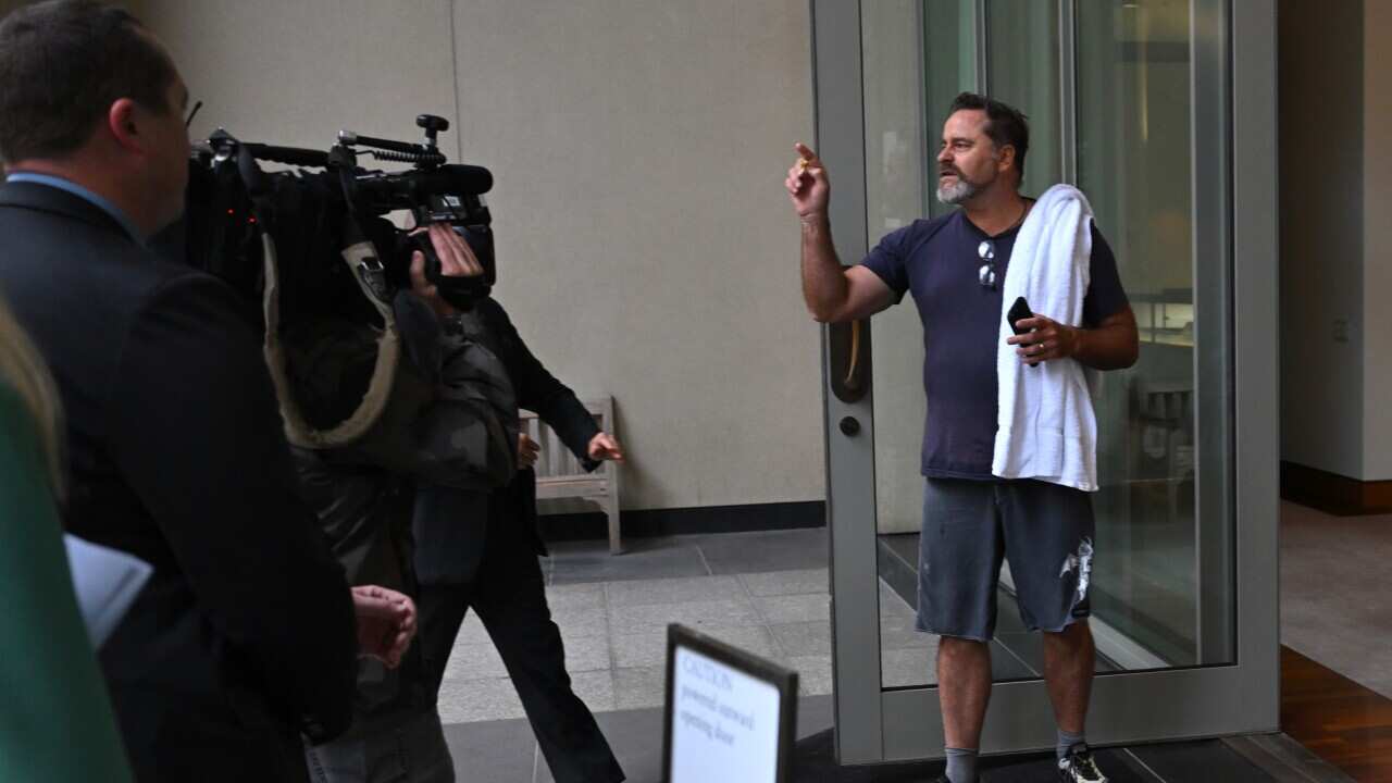 Peter Whish-Wilson, wearing grey shorts and a dark blue t-shirt and with a white towel over his shoulder, yells while standing by a glass door.