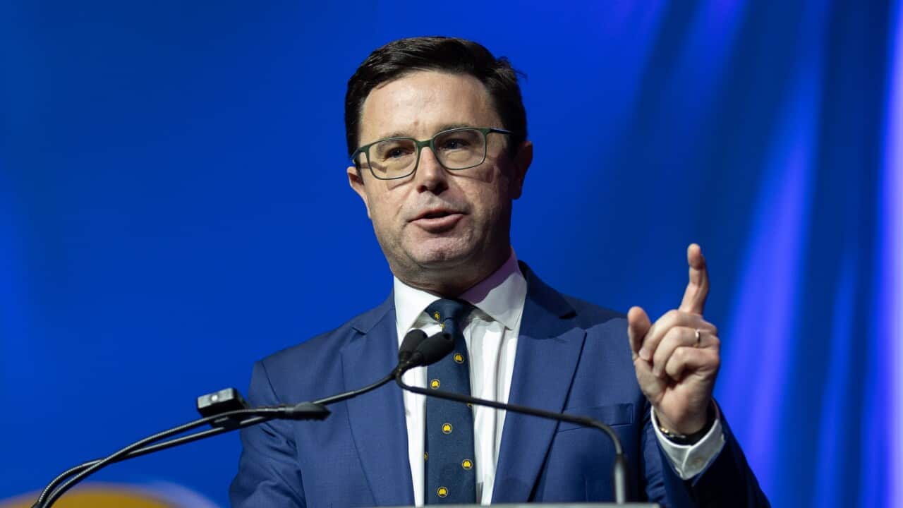 A man in a blue suit raises his finger, pointing upwards as he speaks into microphones in front of a blue background.