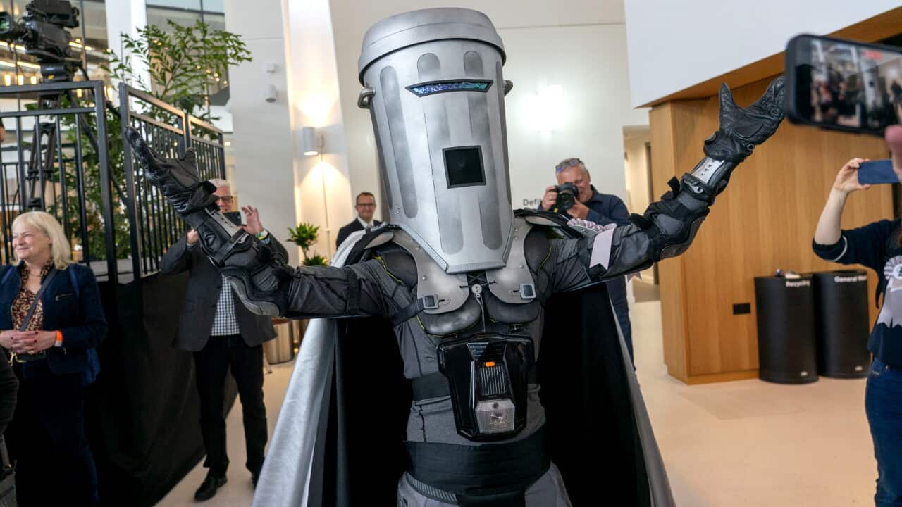A person wearing a silver bin-shaped costume with a cape raises their arms inside a bright building as photographers and onlookers take pictures.
