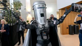 A person wearing a silver bin-shaped costume with a cape raises their arms inside a bright building as photographers and onlookers take pictures.