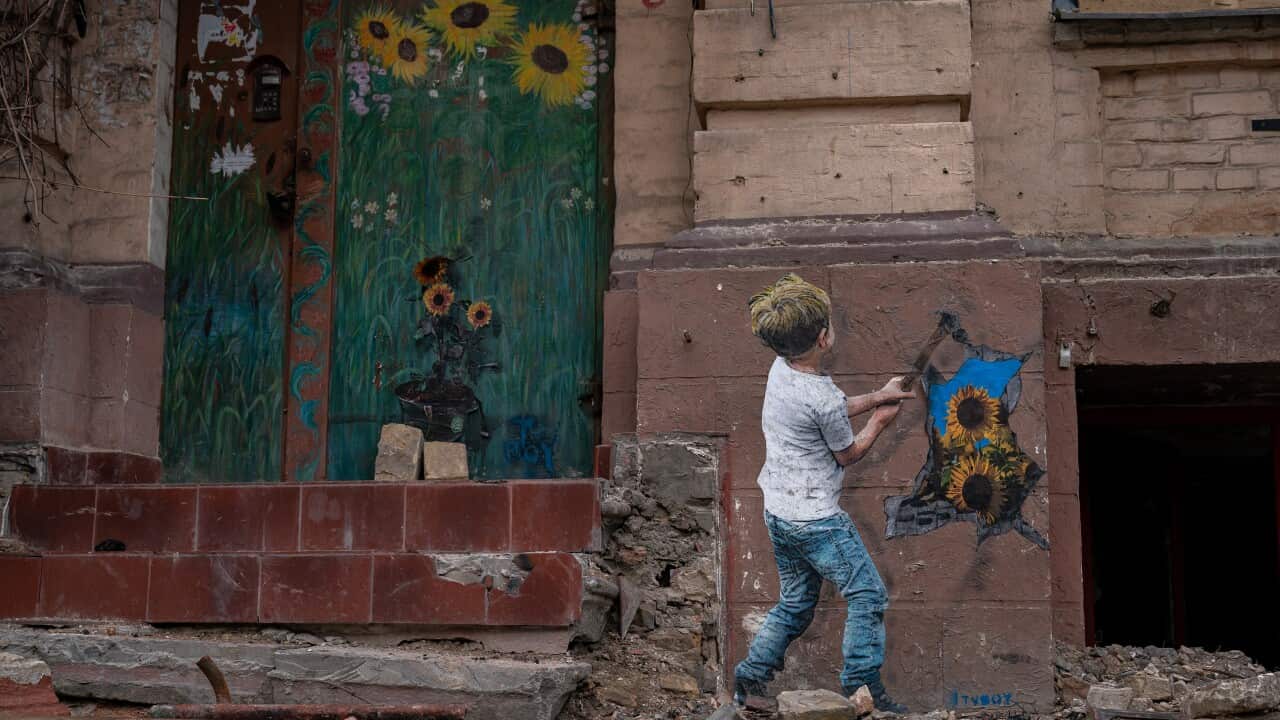 A boy outside a dilapidated building. He is facing the wall. A green wooden door in the building has sunflowers painted on it.