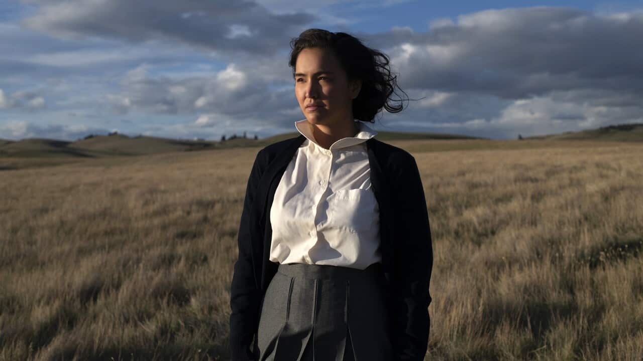 A young woman stands on a grassy plain, with her dark hair blowing in the wind. She wears a white shirt, grey skirt and black cardigan.