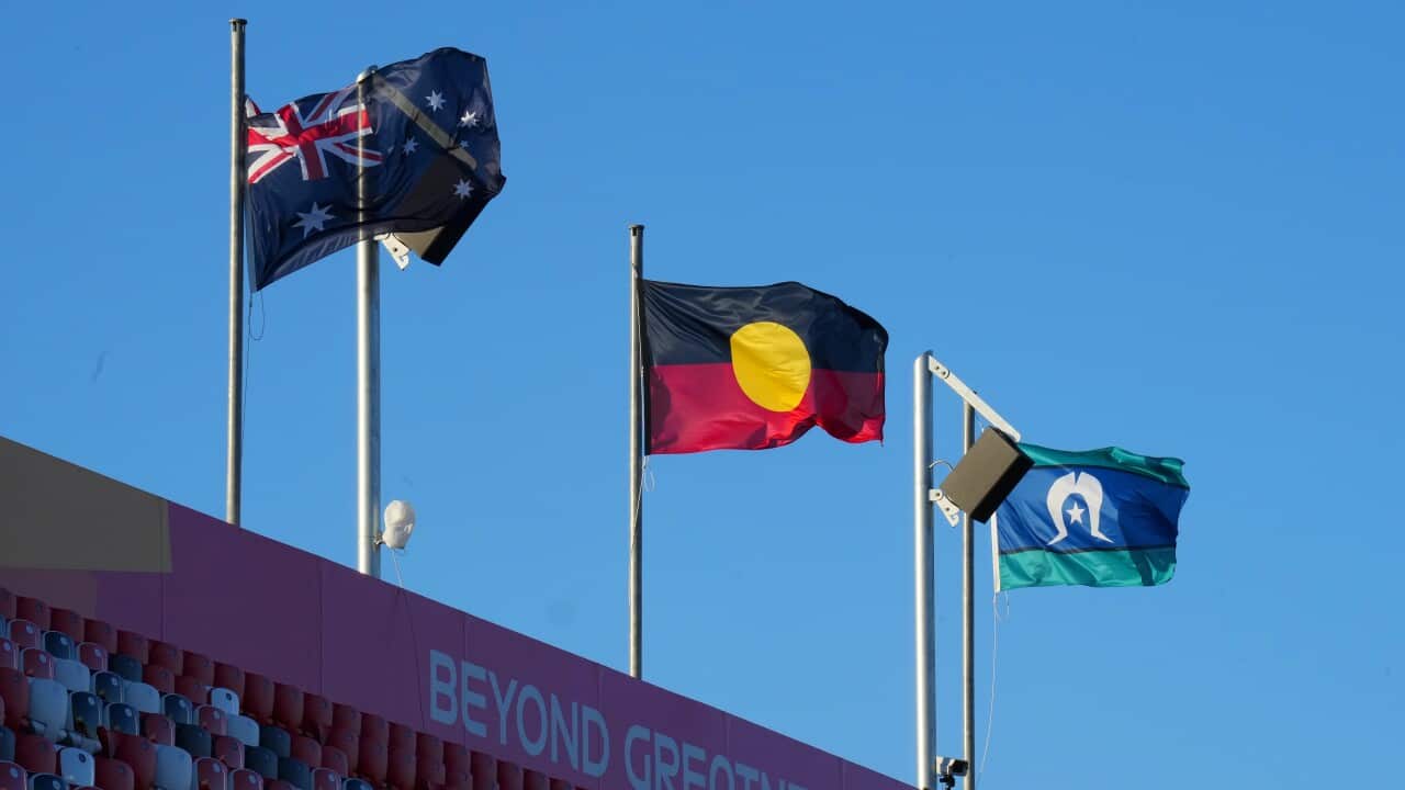 An Australian flag, an Aboriginal flag, and a Torres Strait Island flag are displayed in a row on flagpoles at the top of a stadium.