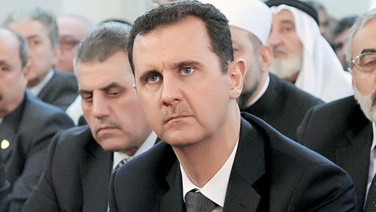 A man in a black suit sits, surrounded by a crowd of men behind him.