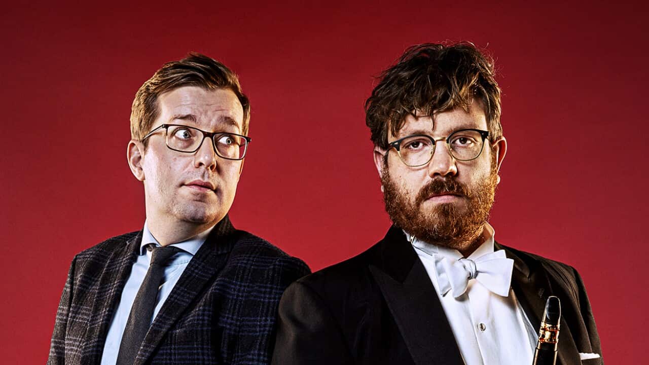 Two men stand together. One, looking straight ahead, is in a formal suit, holding a clarinet. The other looks sideways at the clarinet holder.