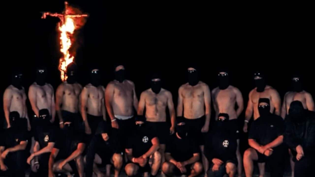 A group of white men stand under a burning cross at Gariwerd, Victoria.