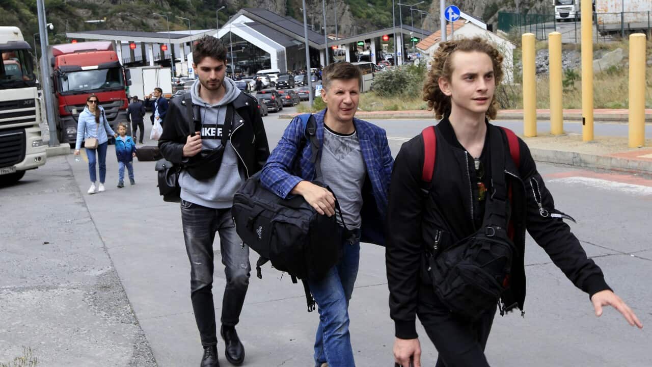 Men walk with backpacks on a road. Behind them is a line of cars.