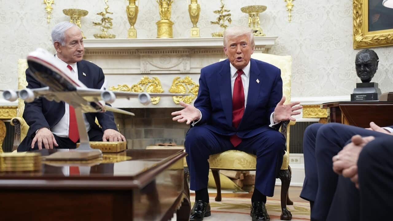 Two men in suits sit on gold chairs as one man speaks, looking directly at the camera.