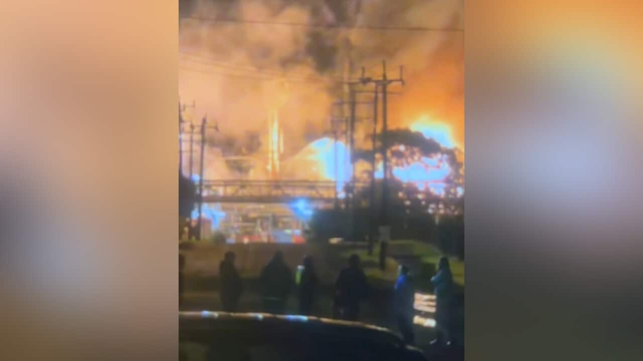 A large, intense industrial fire blazes at night, with bright orange flames and plumes of smoke near power lines and an elevated walkway.