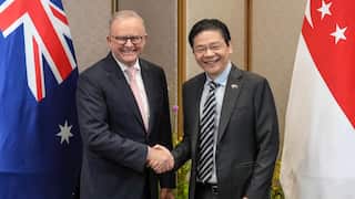 A man in a suit and tie shakes hands with another man in a suit and tie wearing glasses, with Australian and Singaporean flags behind them
