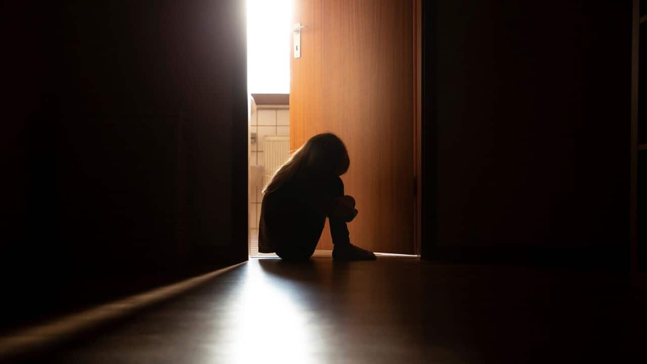 Despairing child sitting with head on knees in the dark frame of a doorway, backlit by a room behind flooded with daylight