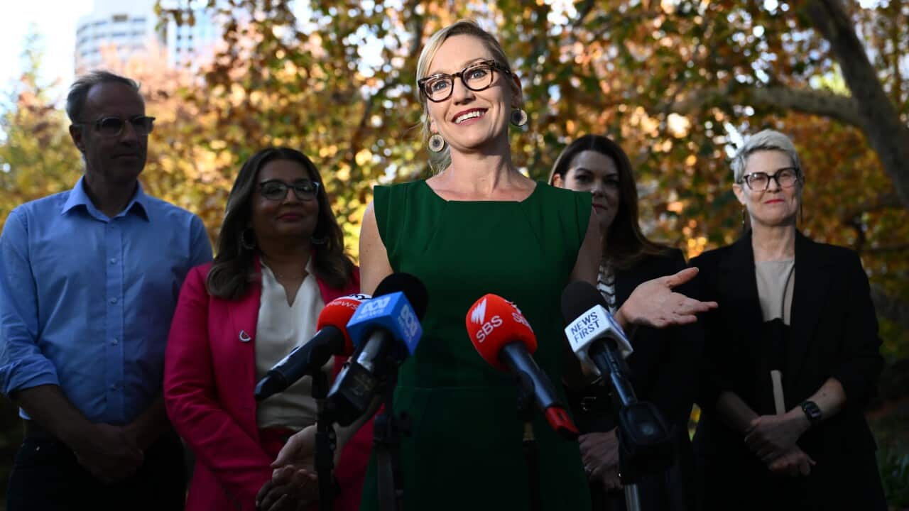 A blonde woman with glasses, speaks in front of media microphones. She is flanked by several other people.