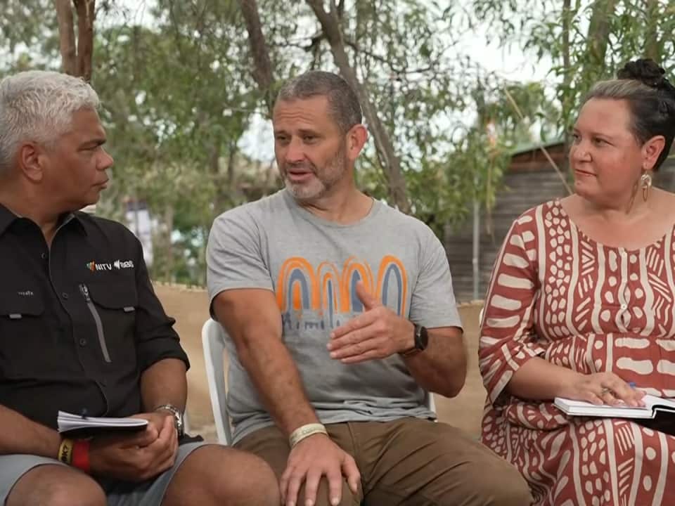 Three people seated outdoors in a panel setting, including an NITV reporter, engaged in an earnest conversation.