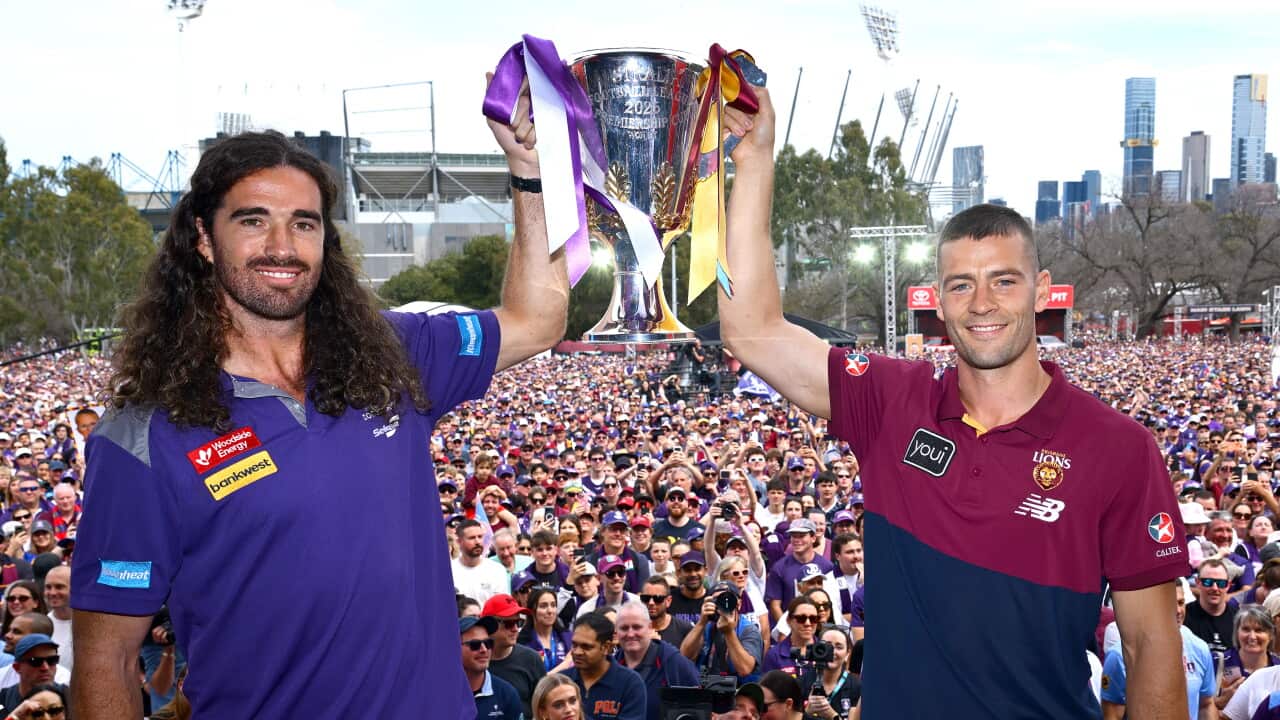 A Fremantle player and a Brisbane Lions player hold the AFL premiership cup above their heads in front of a large crowd in Melbourne.